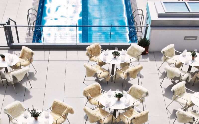 Außenpoolbereich mit runden Tischen und Stühlen, die mit weißem Fell bezogen sind und auf einer gefliesten Terrasse neben einem rechteckigen Swimmingpool stehen.