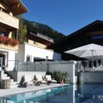 Modernes Gebirgsresort mit Holzbalkonen, Liegestühlen und einem großen weißen Sonnenschirm neben einem klaren Außenpool unter blauem Himmel.