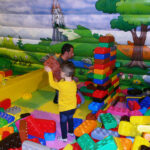 Ein Mann und ein Kind spielen mit großen Plastikbausteinen in einem farbenfrohen Indoor-Spielbereich mit einem Burg-Wandbild an der Wand.