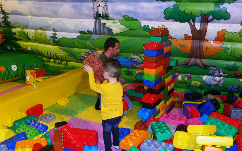Ein Mann und ein Kind spielen mit großen Plastikbausteinen in einem farbenfrohen Indoor-Spielbereich mit einem Burg-Wandbild an der Wand.
