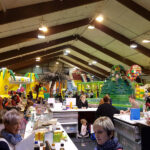 Indoor-Spielplatz mit Hüpfburgen und Rutschen, belebt von Kindern und Erwachsenen. Im Vordergrund befindet sich ein Thekenbereich, in dem Speisen und Waren angeboten werden, und Mitarbeiter, die sich um die Kunden kümmern.