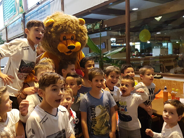 Eine Gruppe Kinder posiert und lächelt mit einer Person in einem Löwenkostüm in einem hell erleuchteten Innenraum.