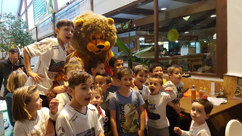 Eine Gruppe Kinder posiert und lächelt mit einer Person in einem Löwenkostüm in einem hell erleuchteten Innenraum.