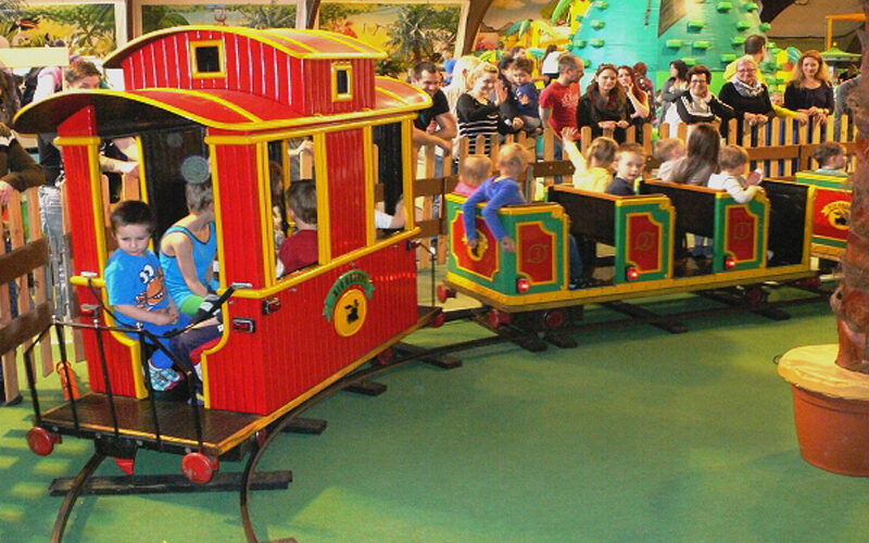 Kinder fahren in einem Vergnügungszentrum mit einer kleinen Indoor-Eisenbahn, während Erwachsene und andere Kinder hinter einem Zaun zuschauen.