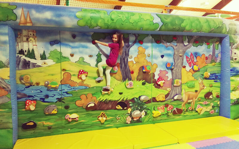 Ein Kind in einem roten Outfit klettert eine bunte Indoor-Kletterwand mit einem Wald- und Burgwandbild über gelben Polstermatten hoch.