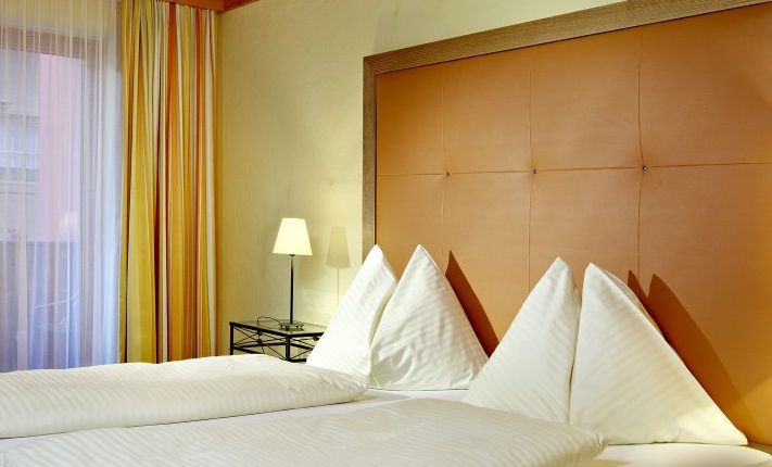 Ein ordentlich gemachtes Hotelbett mit vier dreieckigen weißen Kissen, einem orangefarbenen gepolsterten Kopfteil, einer Nachttischlampe und gestreiften Vorhängen am Fenster.