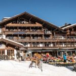Ein hölzernes Alpenresort mit Sitzgelegenheiten im Freien auf einer verschneiten Terrasse; die Menschen speisen und entspannen unter blauem Himmel.