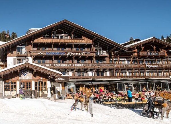 Ein hölzernes Alpenresort mit Sitzgelegenheiten im Freien auf einer verschneiten Terrasse; die Menschen speisen und entspannen unter blauem Himmel.
