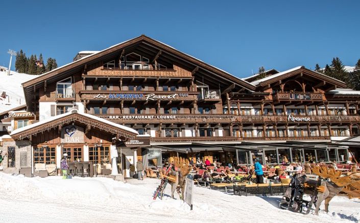 Ein hölzernes Alpenresort mit Sitzgelegenheiten im Freien auf einer verschneiten Terrasse; die Menschen speisen und entspannen unter blauem Himmel.