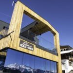 Modernes hölzernes Kurhaus mit großen Glasfenstern, in denen sich schneebedeckte Berge spiegeln, unter einem klaren blauen Himmel. Auf einem Schild steht "Felsen BAD + SPA".