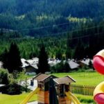 Kinderspielplatz mit Klettergerüsten und einer hohen rot-gelben Spiralrutsche, auf einer Wiese mit Häusern und bewaldeten Hügeln im Hintergrund.