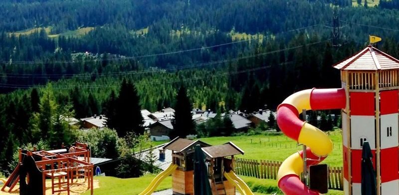 Kinderspielplatz mit Klettergerüsten und einer hohen rot-gelben Spiralrutsche, auf einer Wiese mit Häusern und bewaldeten Hügeln im Hintergrund.