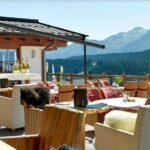 Restaurantterrasse mit gepolsterten Sitzgelegenheiten, gedeckten Tischen, ausgestellten Weinflaschen und einem Blick auf die Berge im Hintergrund.