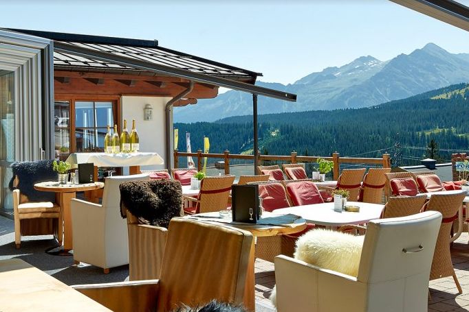 Restaurantterrasse mit gepolsterten Sitzgelegenheiten, gedeckten Tischen, ausgestellten Weinflaschen und einem Blick auf die Berge im Hintergrund.