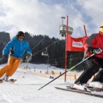 Zwei Personen fahren auf einer verschneiten Piste mit Skiliften und Kiefern im Hintergrund bergab; die eine trägt rot, die andere blau und orange.