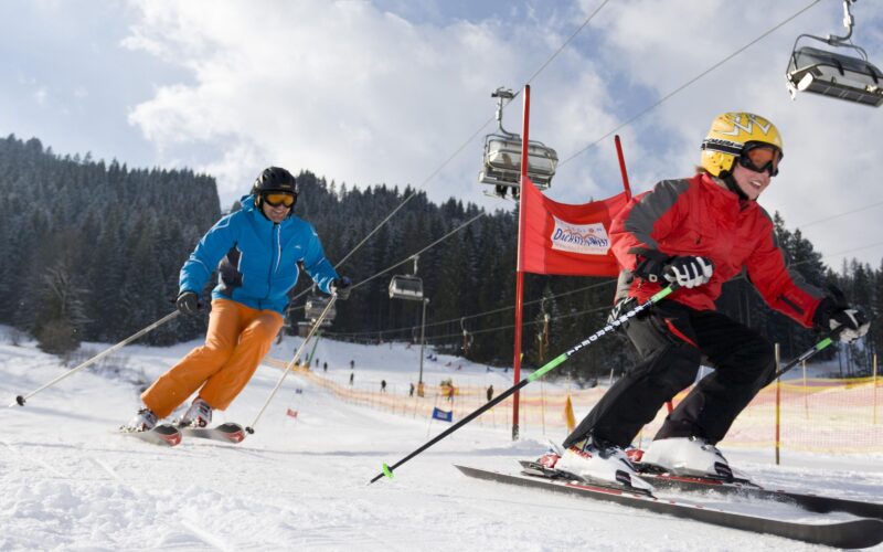 Zwei Personen fahren auf einer verschneiten Piste mit Skiliften und Kiefern im Hintergrund bergab; die eine trägt rot, die andere blau und orange.