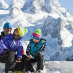 Eine vierköpfige, winterlich gekleidete Familie sitzt und steht mit Skiern im Schnee, umgeben von schneebedeckten Bergen im Hintergrund.