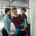 Ein Mann hält einen lächelnden kleinen Jungen, während er neben einer Frau steht; alle drei schauen auf eine Anzeige an der Wand in einem hellen Innenraum.