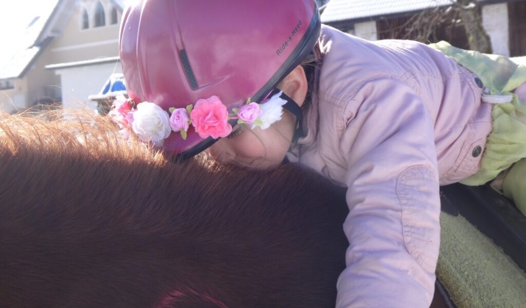Ein Kind mit rosa Helm und Blumenkrone umarmt ein braunes Pferd an einem sonnigen Tag im Freien, mit Häusern im Hintergrund.