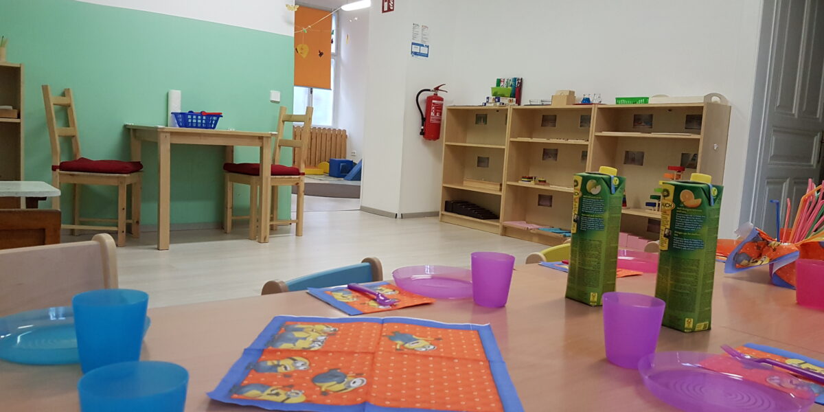 Ein hell erleuchtetes Klassenzimmer mit Tischen, die für eine Mahlzeit gedeckt sind, mit bunten Tellern, Tassen, Saftkartons und Servietten, mit Regalen, Stühlen und pädagogischem Dekor im Hintergrund.