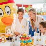 Kinder und ein Erwachsener versammeln sich um eine Geburtstagstorte mit Wunderkerzen; eine Person in einem großen gelben Vogelkostüm steht hinter ihnen. Auf dem Tisch sind Partydekorationen und Hüte zu sehen.
