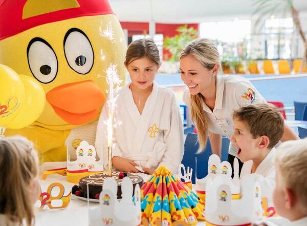 Kinder und ein Erwachsener versammeln sich um eine Geburtstagstorte mit Wunderkerzen; eine Person in einem großen gelben Vogelkostüm steht hinter ihnen. Auf dem Tisch sind Partydekorationen und Hüte zu sehen.