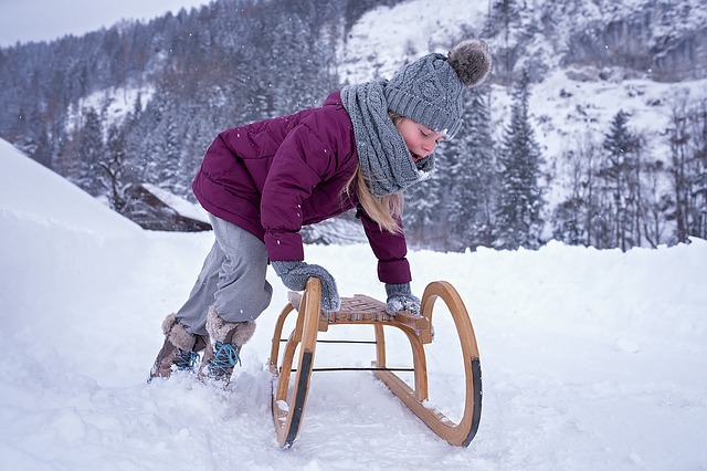 Ein Kind in Winterkleidung beugt sich vor, um einen Holzschlitten durch den Schnee zu schieben, mit verschneiten Bäumen und Bergen im Hintergrund.
