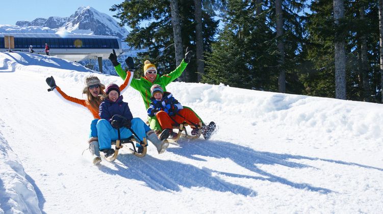 Zwei Erwachsene und zwei Kinder rodeln einen verschneiten Hügel hinunter, mit Bäumen und Bergen im Hintergrund, alle tragen Winterkleidung und lächeln.