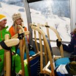 Vier Personen in Winterkleidung sitzen in einer Skigondel mit Holzschlitten, durch die Fenster sind verschneite Berge zu sehen.