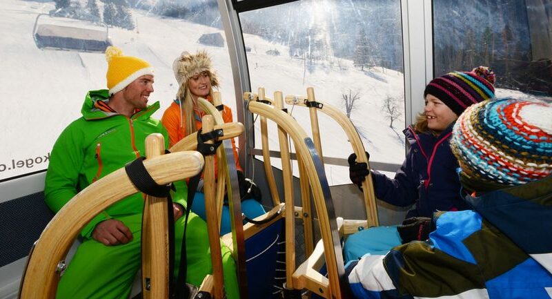 Vier Personen in Winterkleidung sitzen in einer Skigondel mit Holzschlitten, durch die Fenster sind verschneite Berge zu sehen.