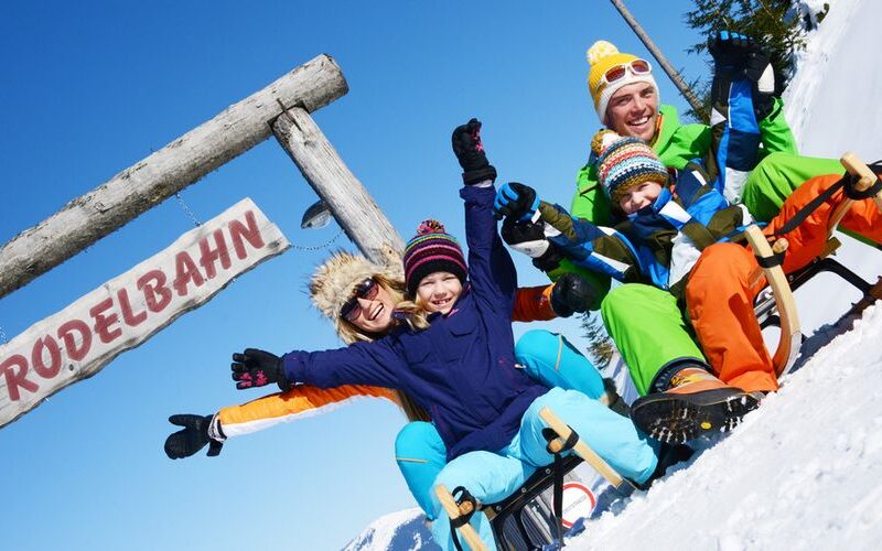Eine winterlich gekleidete Familie sitzt auf Schlitten unter einem Holzschild mit der Aufschrift "RODELBAHN", lächelt und hebt an einem sonnigen Tag die Arme in den Schnee.