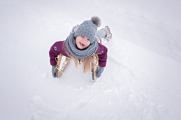 Ein winterlich gekleidetes Kind liegt mit dem Gesicht nach unten auf einem Holzschlitten im Schnee, schaut nach oben und lächelt.