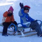 Zwei Kinder mit Winterjacken und Mützen sitzen auf Schlitten im Schnee und winken der Kamera zu.