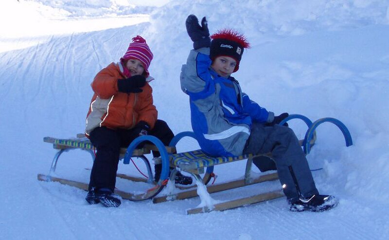 Zwei Kinder mit Winterjacken und Mützen sitzen auf Schlitten im Schnee und winken der Kamera zu.