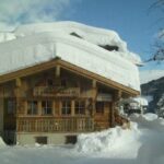Ein Holzchalet mit einer dicken Schneeschicht auf dem Dach und dem umliegenden Boden, eingebettet in eine verschneite Berglandschaft unter blauem Himmel.