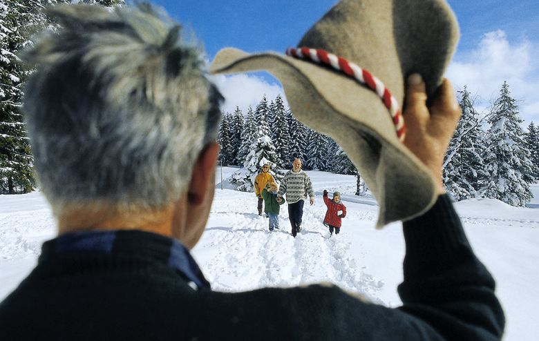 Eine Person mit Hut steht vor einer Gruppe von drei Personen, darunter ein Kind, die in einer verschneiten Landschaft mit Bäumen im Hintergrund auf sie zugehen.
