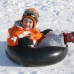 Ein kleines Kind in Winterkleidung sitzt auf einem schwarzen Fahrradschlauch im Schnee, lächelt und lehnt sich mit ausgestreckten Beinen zurück.