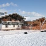 Eine Holzhütte und ein Nebengebäude sind von Schnee umgeben, im Vordergrund befinden sich ein Maschendrahtzaun und verstreute Reifen; im Hintergrund sind Berge zu sehen.