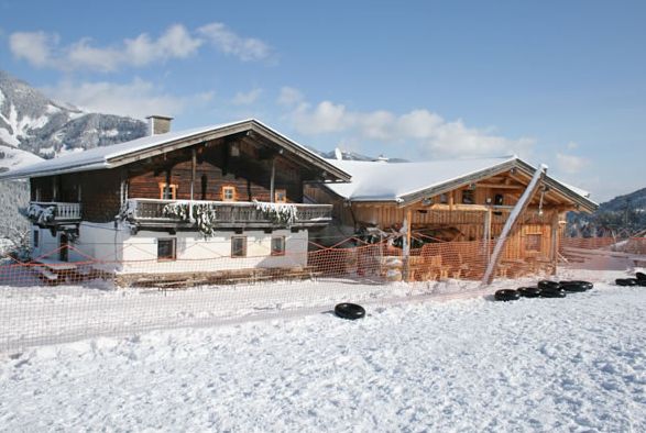 Eine Holzhütte und ein Nebengebäude sind von Schnee umgeben, im Vordergrund befinden sich ein Maschendrahtzaun und verstreute Reifen; im Hintergrund sind Berge zu sehen.