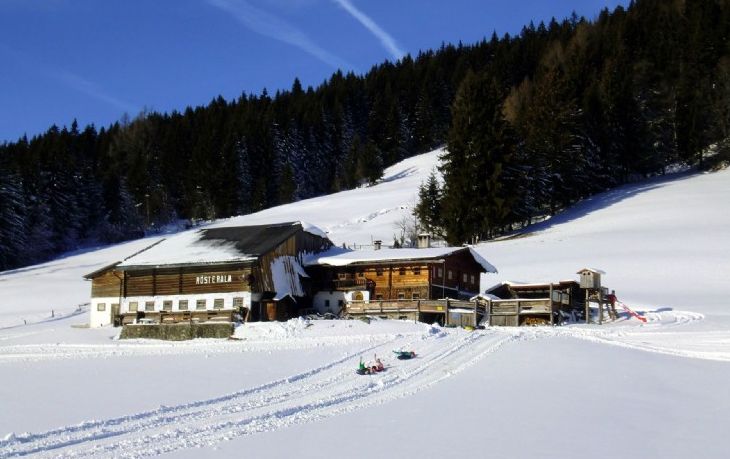 Eine rustikale Holzhütte liegt neben einem schneebedeckten Feld mit Schlittenspuren, umgeben von einem dichten, immergrünen Wald unter einem strahlend blauen Himmel.