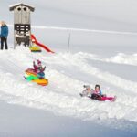 Kinder rutschen auf aufblasbaren Schlitten neben einer kleinen Holzkonstruktion die verschneiten Wege hinunter, während ein Erwachsener in der Nähe steht und zusieht.