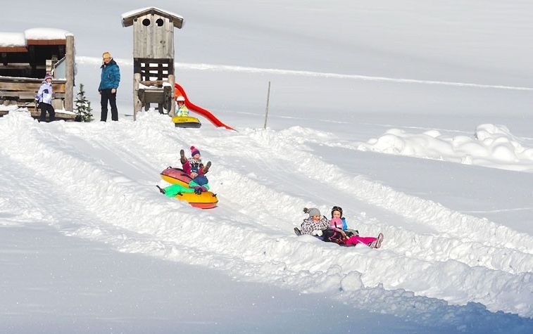 Kinder rutschen auf aufblasbaren Schlitten neben einer kleinen Holzkonstruktion die verschneiten Wege hinunter, während ein Erwachsener in der Nähe steht und zusieht.