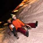 Zwei Personen in Winterkleidung fahren nachts mit einem Schlitten einen verschneiten Hügel hinunter, wobei verschwommene Lichter im Hintergrund die Bewegung anzeigen.