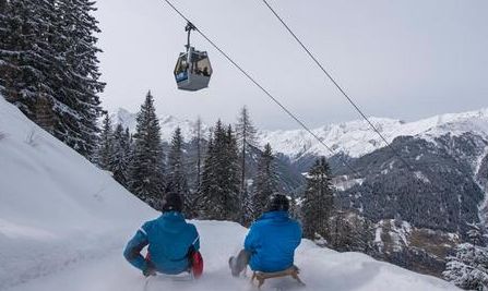 Zwei Personen in Winterjacken rodeln einen verschneiten Hügel hinunter, während über ihnen eine Seilbahn vorbeifährt und im Hintergrund schneebedeckte Berge zu sehen sind.