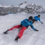 Zwei Personen in Winterkleidung fahren mit Schlitten einen verschneiten Hang hinunter, mit Bergen im Hintergrund.