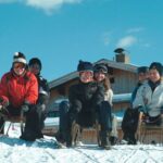 Fünf Personen in Winterkleidung sitzen auf Schlitten auf einem verschneiten Hügel vor einer Holzhütte unter blauem Himmel.