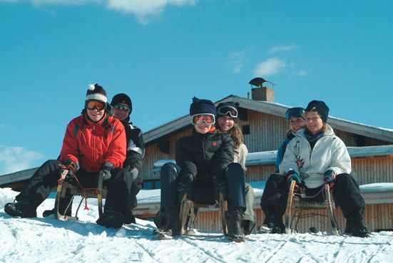 Fünf Personen in Winterkleidung sitzen auf Schlitten auf einem verschneiten Hügel vor einer Holzhütte unter blauem Himmel.