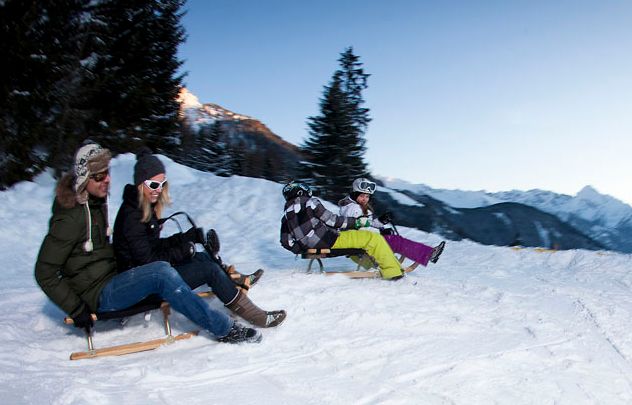 Drei Personen fahren auf Holzschlitten einen verschneiten Hang hinunter, im Hintergrund sind schneebedeckte Bäume und Berge zu sehen.