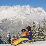 Zwei Personen mit Helmen und Winterkleidung schlittern einen verschneiten Hügel hinunter, mit Kiefern und schneebedeckten Bergen im Hintergrund.