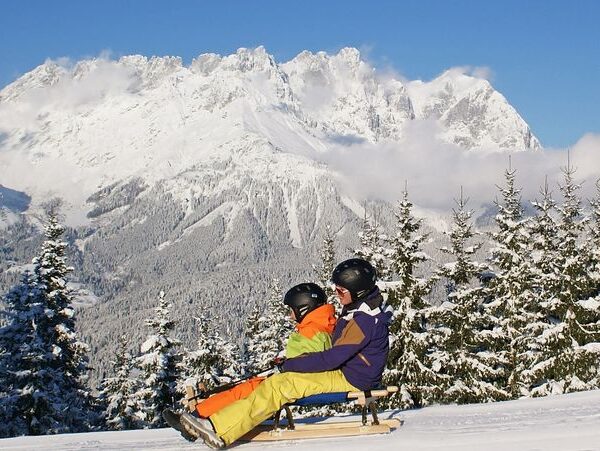 Zwei Personen mit Helmen und Winterkleidung schlittern einen verschneiten Hügel hinunter, mit Kiefern und schneebedeckten Bergen im Hintergrund.